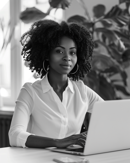 Black and white image of a confident African American woman entrepreneur working at a laptop in a minimalist home office, symbolizing female empowerment and digital business success. www.elevatehervault.com